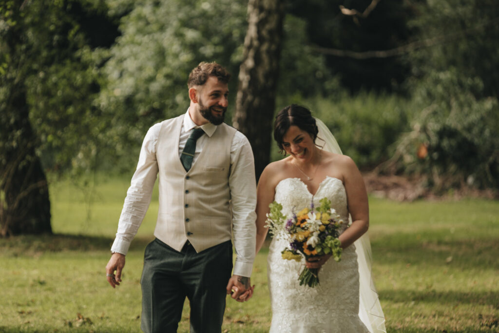 A smiling couple walks hand in hand outdoors on grass. The man wears a beige vest, white shirt, and tie, while the woman in a white lace wedding dress holds a bouquet. Trees and blurred greenery are in the background, suggesting a forest or garden setting. © Aimee Lince Photography at The Dower House Hotel