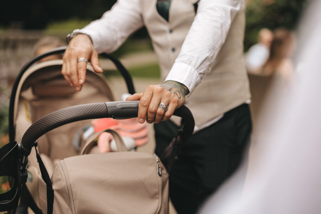 A person wearing a white shirt and beige vest with tattooed arms gently holds a stroller handle. The stroller is beige, and a pink object is visible inside. The background is blurred, with hints of greenery and people, suggesting an outdoor setting. © Aimee Lince Photography at The Dower House Hotel