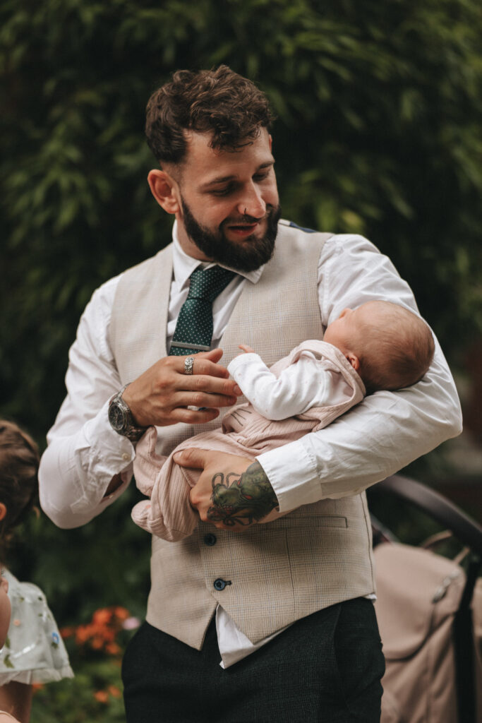 A bearded man with a tattoo on his forearm holds a baby. He wears a white shirt, beige vest, and a green tie, smiling down at the infant dressed in a pale pink outfit. They stand against a backdrop of greenery, with a stroller partially visible. © Aimee Lince Photography at The Dower House Hotel