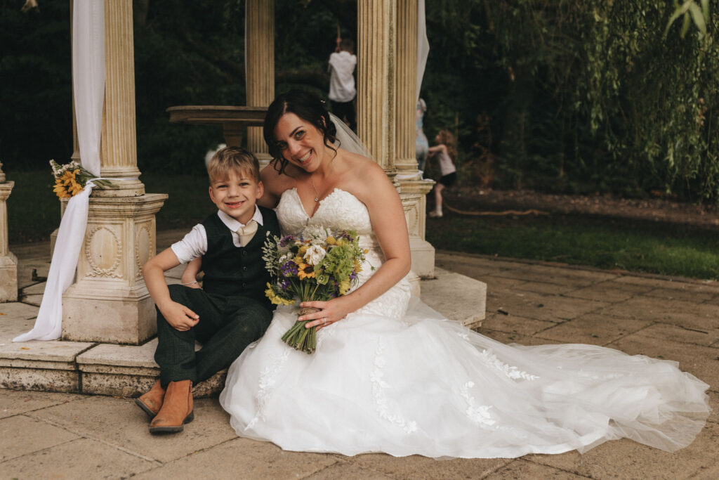 A bride in a white strapless gown sits beside a young boy on a stone pathway. She holds a bouquet of wildflowers and smiles warmly. The boy, dressed in a vest, shirt, and boots, sits close to her. They are in front of a columned structure with draped fabric, surrounded by lush greenery. © Aimee Lince Photography at The Dower House Hotel