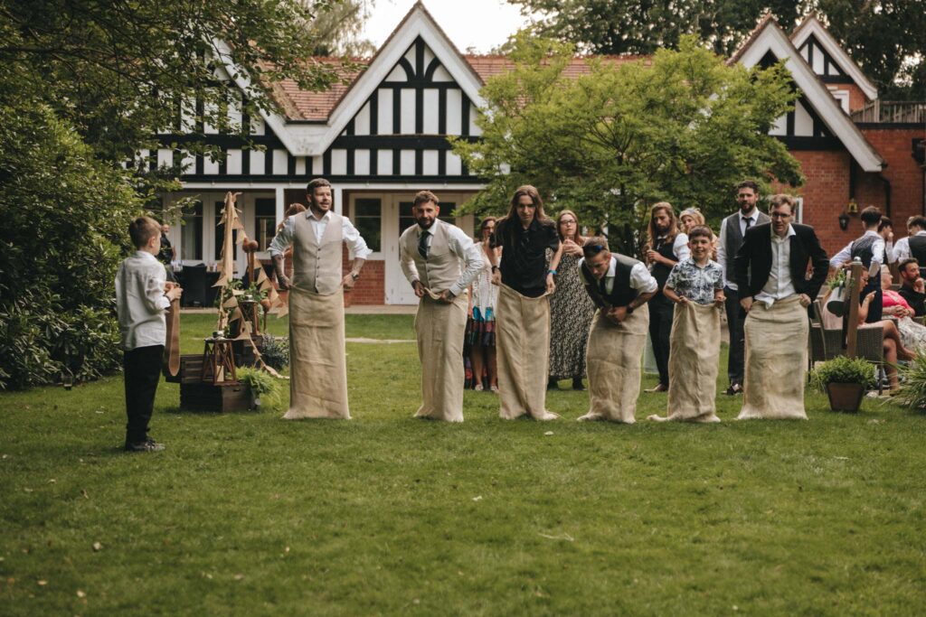 People are participating in a sack race on a lush green lawn in front of a Tudor-style house. Five adults in mid-jump appear focused and competitive. Spectators are gathered around, watching the event. Trees and shrubs border the garden area, enhancing the lively outdoor atmosphere. © Aimee Lince Photography at The Dower House Hotel