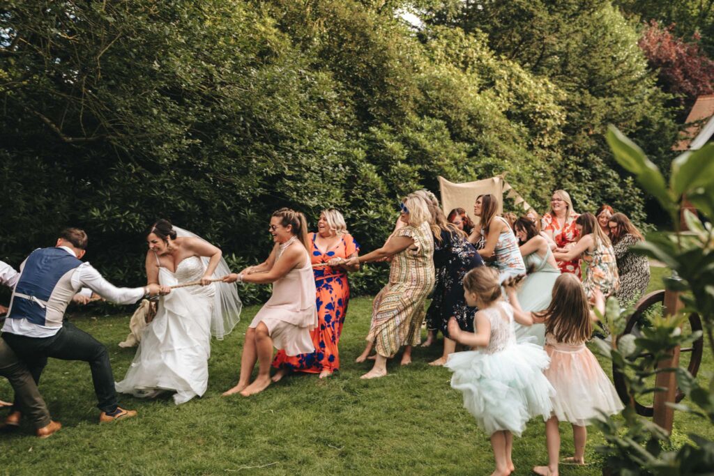 A lively outdoor tug-of-war between men and women at a wedding. The bride, in a white gown, leads the women's team on grassy ground surrounded by lush greenery. Guests, including children in pastel dresses, cheer and participate energetically under sunlight. © Aimee Lince Photography at The Dower House Hotel
