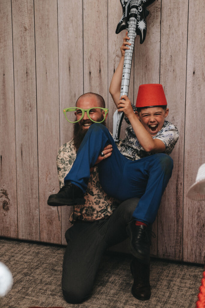 A bearded man and a boy pose playfully. The man wears oversized green glasses and holds the boy aloft. The boy, wearing a red fez and blue pants, holds an inflatable guitar. They are in front of a wooden backdrop, both smiling energetically. © Aimee Lince Photography at The Dower House Hotel
