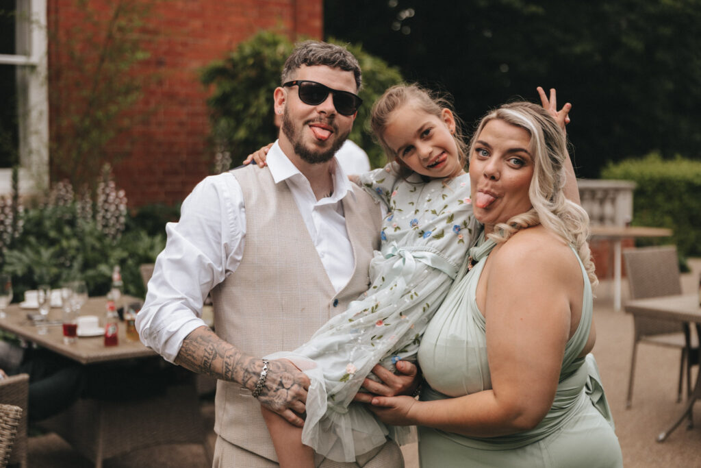 A man, woman, and young girl pose playfully for the camera, sticking out their tongues. The man wears sunglasses, a white shirt, and a beige vest. The woman wears a light green dress, and the girl wears a floral dress. Patio furniture and greenery are visible in the background. © Aimee Lince Photography at The Dower House Hotel