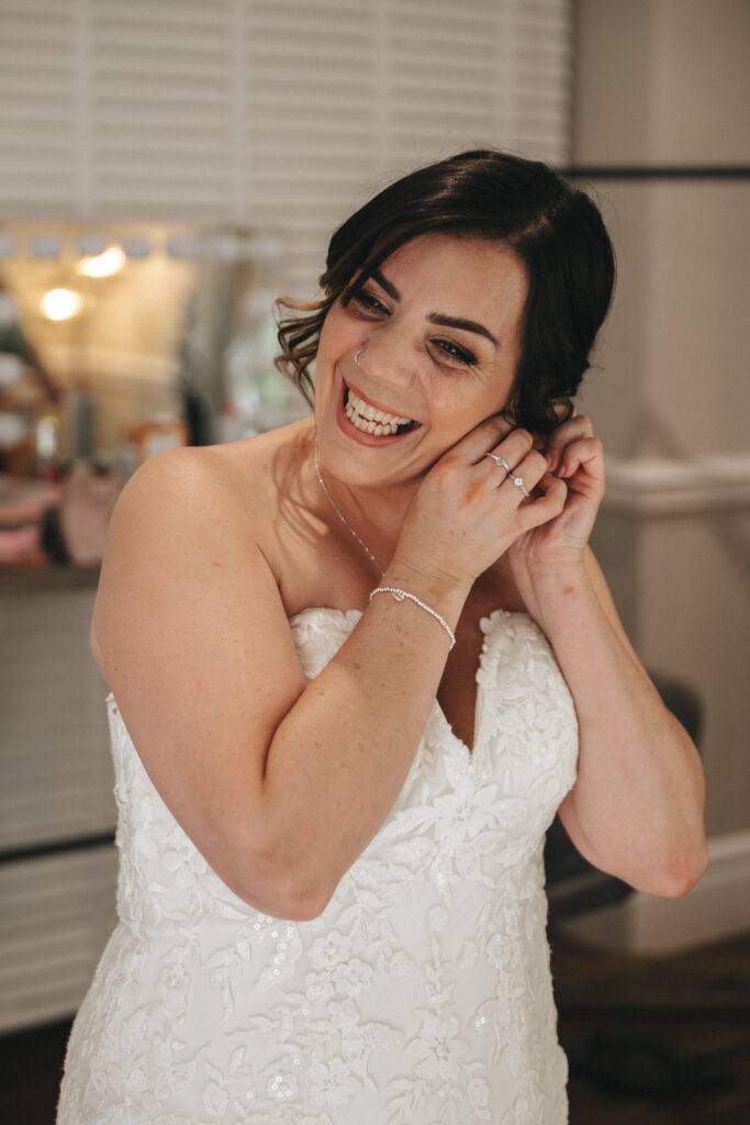 A woman with dark hair smiles as she puts on an earring. She is wearing a strapless white lace wedding dress and a delicate necklace. The room has soft lighting, with a dressing table and mirror in the background, creating an intimate, joyful atmosphere. © Aimee Lince Photography at The Dower House Hotel