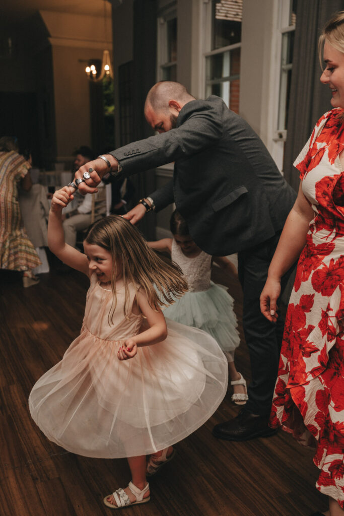 A man in a gray suit jacket twirls a young girl wearing a light pink dress and white sandals on a wooden floor. Another child in a green dress is in the background. A woman in a red and white floral dress smiles nearby. The setting appears to be an indoor event with dim lighting. © Aimee Lince Photography at The Dower House Hotel