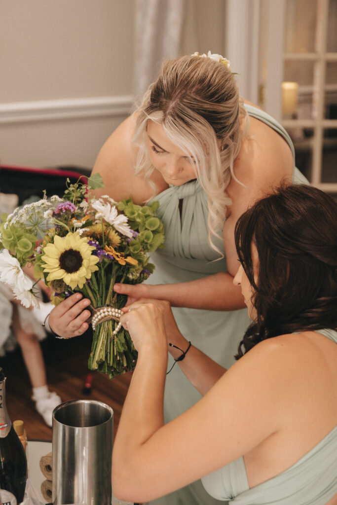 Two women in turquoise dresses arranging a bouquet of sunflowers and white flowers. One woman is holding the bouquet, while the other adjusts a pearl bracelet around the stems. They are in a room with wooden flooring and soft lighting, and a girl is visible in the background. © Aimee Lince Photography at The Dower House Hotel