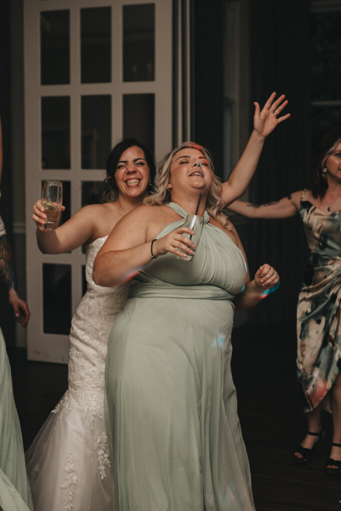 Two women celebrate at a party. The woman in a white wedding dress holds a drink and smiles while standing behind another woman in a mint green dress. The woman in green raises her hand with eyes closed in a joyful gesture. Other guests are partially visible in the background. © Aimee Lince Photography at The Dower House Hotel