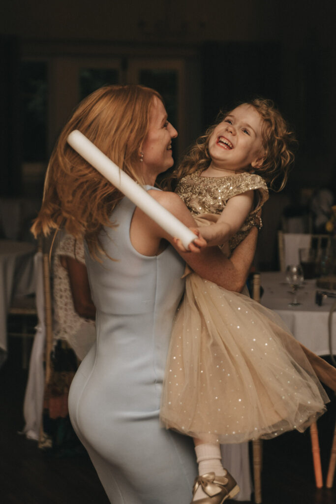 A woman in a light blue dress joyfully spins with a young girl wearing a sparkly gold dress. The girl holds a white stick and laughs. The background features blurred dining tables and chairs, suggesting a festive or celebratory atmosphere. © Aimee Lince Photography at The Dower House Hotel