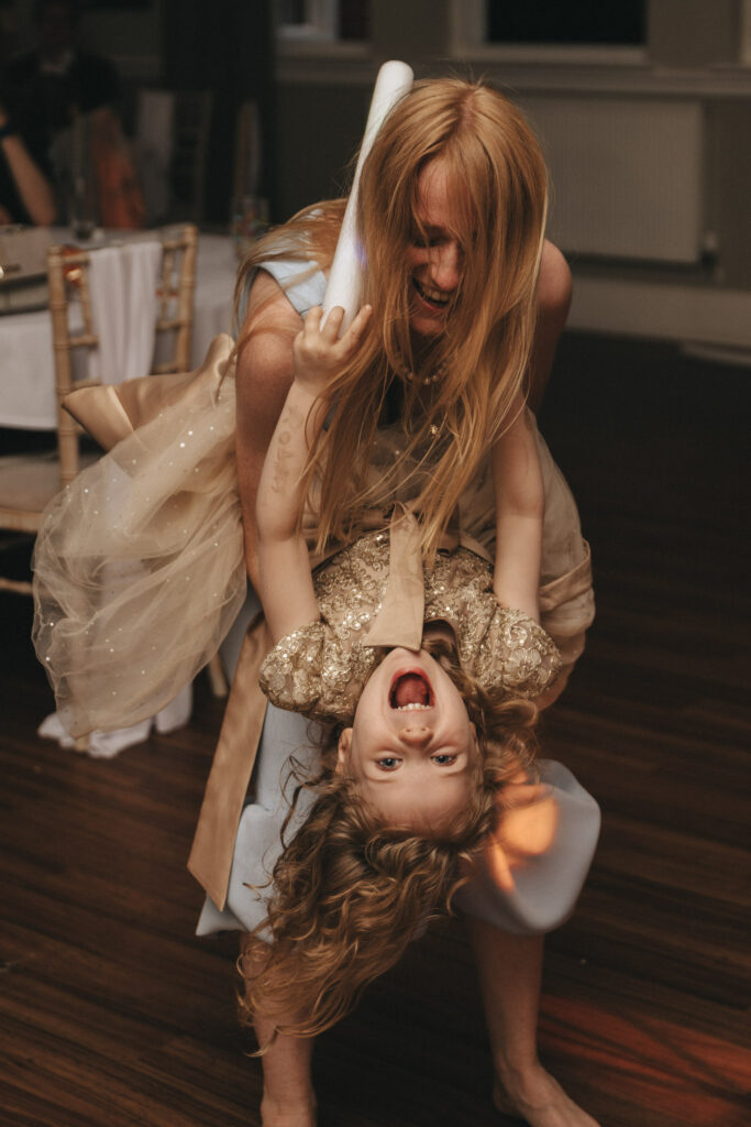 A woman playfully holds an upside-down girl with curly hair, both laughing joyfully. The woman wears a beige dress, and the girl is in a sparkly gold dress. They are indoors, with wooden flooring and a table with a white tablecloth visible in the background. The scene is lively and joyful. © Aimee Lince Photography at The Dower House Hotel