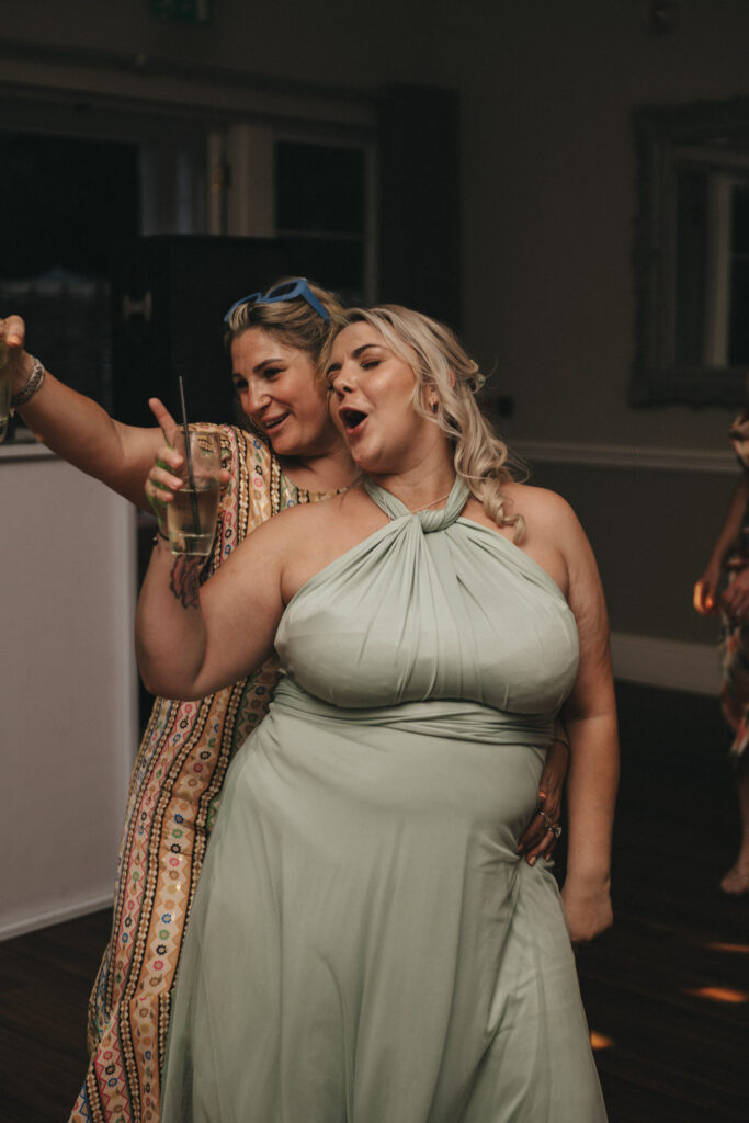 Two women smiling and dancing together at a party. The one on the left is wearing a colorful patterned dress and holding a drink, while the other, with blonde hair, is in a light green, halter-neck dress. They appear joyful and are positioned in front of a plain wall. © Aimee Lince Photography at The Dower House Hotel