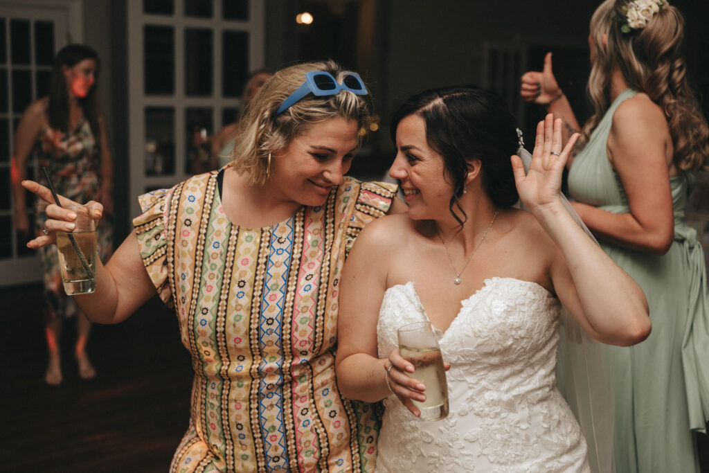Dower House Hotel Wedding - Lincolnshire Photographer - Charlotte & Kris © Aimee Lince Photography (628) Two women dance and smile at an indoor celebration. The woman on the left wears a colorful patterned dress and holds a drink, with sunglasses on her head. The woman on the right is in a white strapless dress, also holding a drink. A bridesmaid in green is visible in the background. © Aimee Lince Photography at The Dower House Hotel