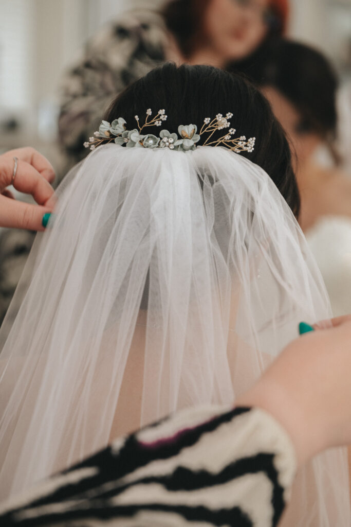 A bride getting ready, with hands adjusting a white veil adorned with small white and blue flowers and gold accents. The background is softly blurred, showing someone in a patterned outfit. The bride's dark hair is styled up beneath the veil. © Aimee Lince Photography at The Dower House Hotel