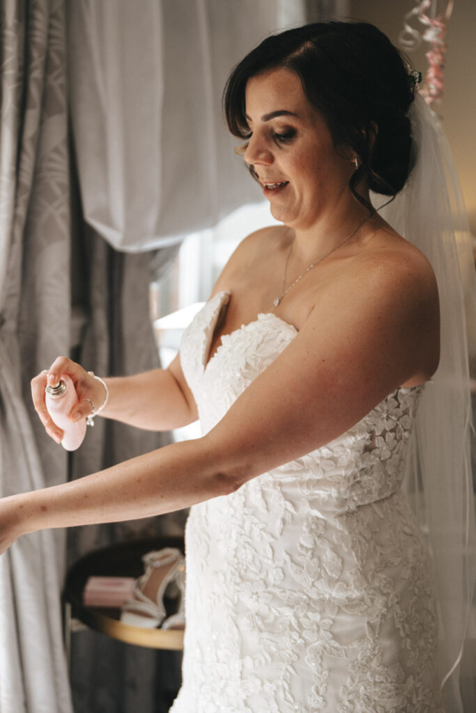 A bride in a white lace strapless gown sprays perfume on her wrist. She wears a veil and a bracelet, standing near a window with sheer curtains. A small table with shoes and a pink box is in the background. Her dark hair is styled in an updo. © Aimee Lince Photography at The Dower House Hotel