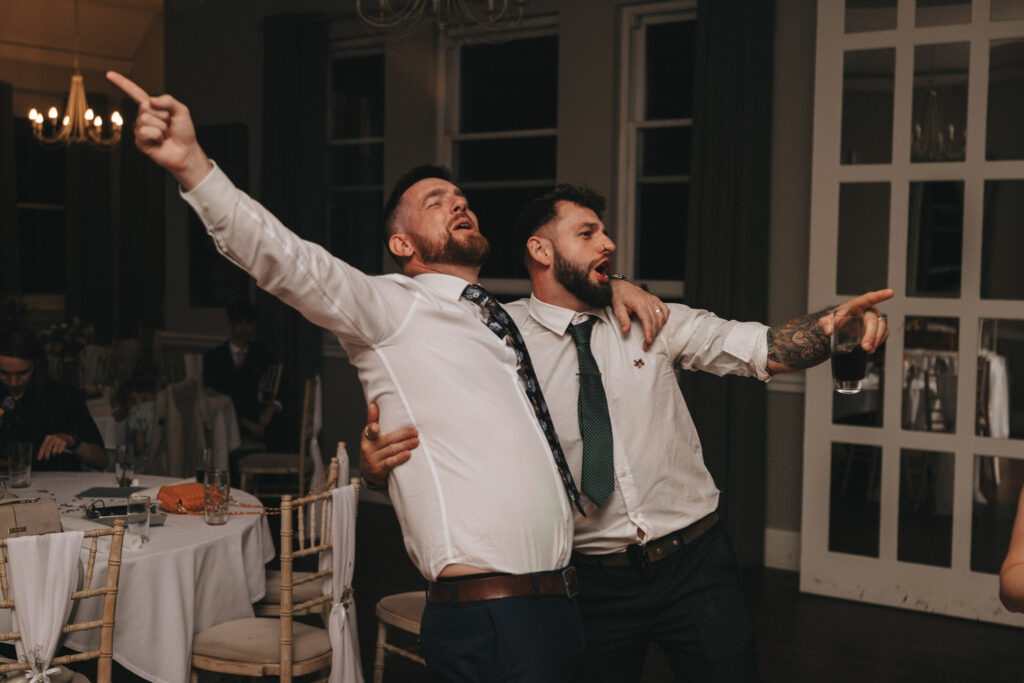 Two men in white shirts and ties enthusiastically sing while holding each other, one with a drink. They're in a dimly lit room with round tables and white chairs in the background. The mood is joyful, suggesting a celebration or party atmosphere. © Aimee Lince Photography at The Dower House Hotel