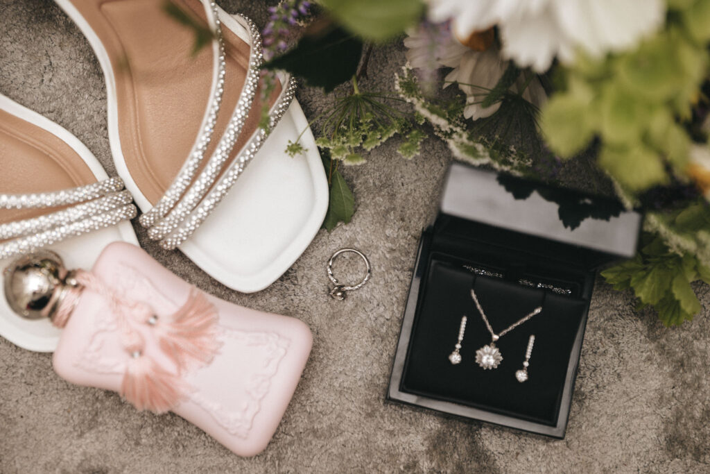 Elegant flat lay of bridal accessories on a textured surface: white, bejeweled open-toe heels, a delicate ring, a pink ornate perfume bottle, and a black jewelry box containing a matching diamond necklace and earrings set, all accented by lush green and white flowers. © Aimee Lince Photography at The Dower House Hotel
