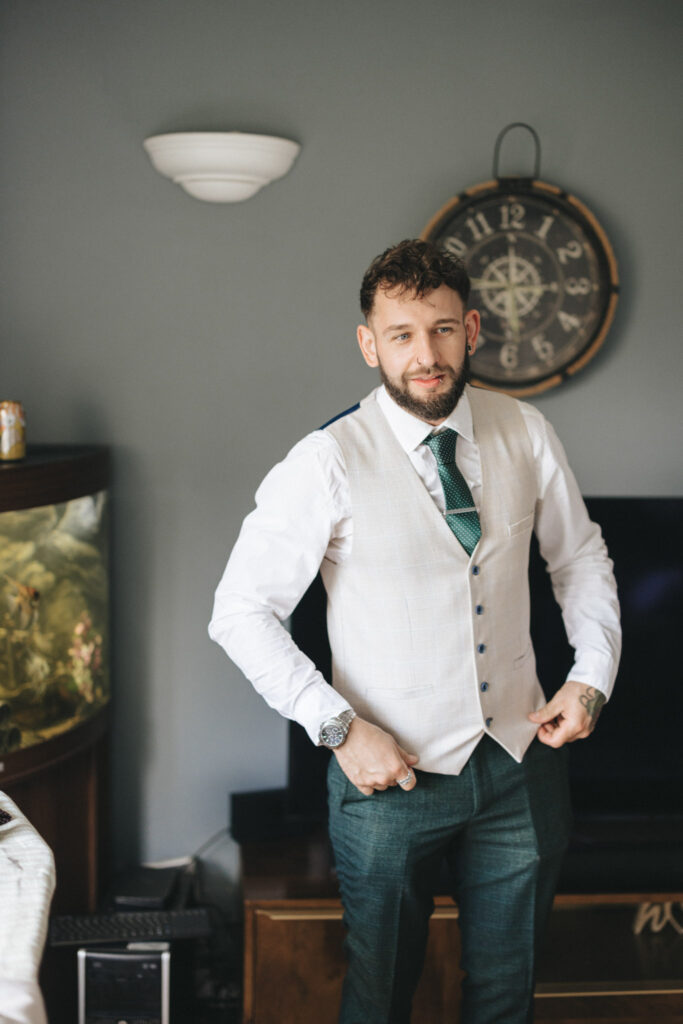 Bearded man in a white shirt, beige vest, and green tie stands in a room with a nautical-themed clock on the wall. He is adjusting his vest. There's a fish tank and a TV behind him. The room has a grey wall and ambient lighting. © Aimee Lince Photography