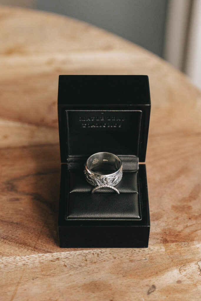 A silver ring with leaf engravings is displayed in an open black velvet ring box. The box is set on a wooden surface. The ring has intricate detailing, and the box's interior is cushioned, enhancing the ring's elegant presentation. © Aimee Lince Photography
