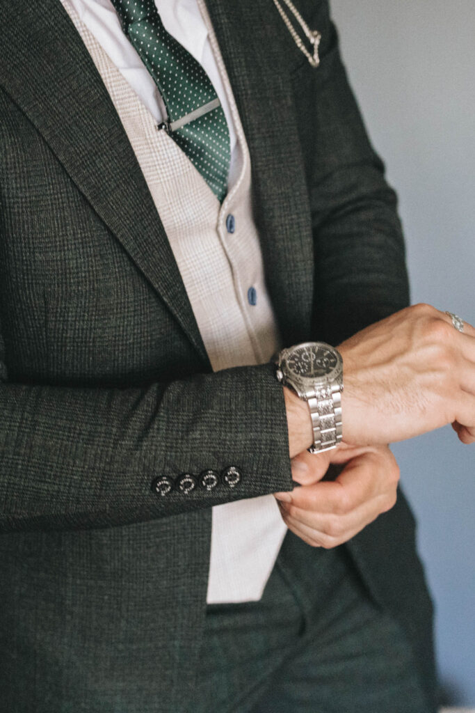 Man in a dark textured suit adjusts his silver wristwatch. He wears a light-colored vest and a green tie with white polka dots. The suit jacket features decorative sleeve buttons. A pocket watch chain is visible under the jacket, highlighting a classic and sophisticated style. © Aimee Lince Photography