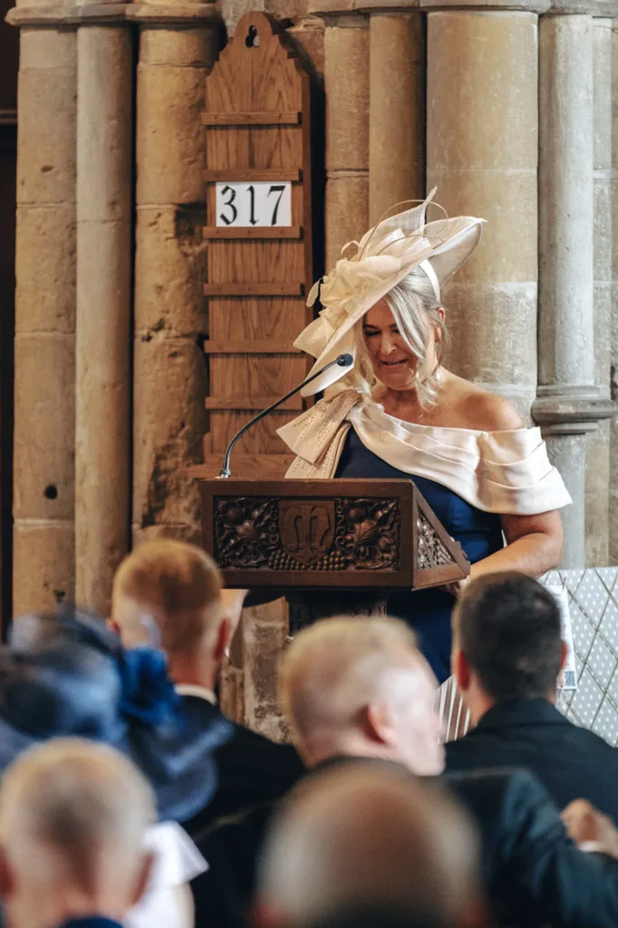 A woman in an off-the-shoulder dress and elaborate fascinator stands at a carved wooden lectern, possibly sharing wedding day tips for the bride and her mum. She addresses an audience inside a stone building, with a wooden sign displaying "317" visible on the wall behind her. © Aimee Lince Photography - Wedding photographer in Lincolnshire, Yorkshire & Nottinghamshire