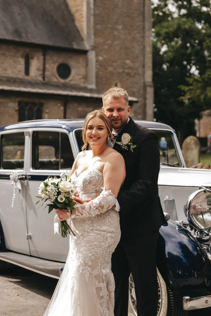 A bride and groom pose elegantly in front of a vintage car, captured with stunning photography. The bride wears a white lace gown, holding a bouquet of white flowers, while the groom dons a black suit with a boutonniere. A half-day package perfectly frames this idyllic scene against the backdrop of stone buildings and trees. © Aimee Lince Photography - Wedding photographer in Lincolnshire, Yorkshire & Nottinghamshire