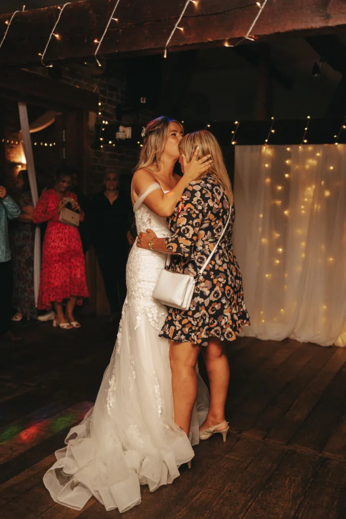 Two women embrace in a warmly lit room adorned with string lights, capturing the essence of mother-daughter moments on a wedding day. The woman on the left wears a white wedding dress, while the other dons a floral dress and carries a white purse. Guests in colorful outfits stand amidst sheer curtains that add ambiance. © Aimee Lince Photography - Wedding photographer in Lincolnshire, Yorkshire & Nottinghamshire