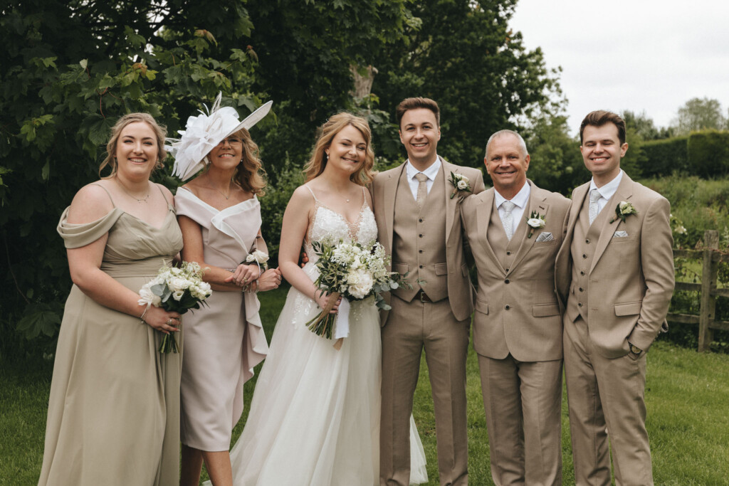 A bride and groom stand with four wedding guests in a garden. The bride wears a white gown and holds a bouquet. The groom and two men wear beige suits, while two women wear light dresses, one with a stylish hat. They all smile, posing in front of trees and greenery. © Aimee Lince Photography