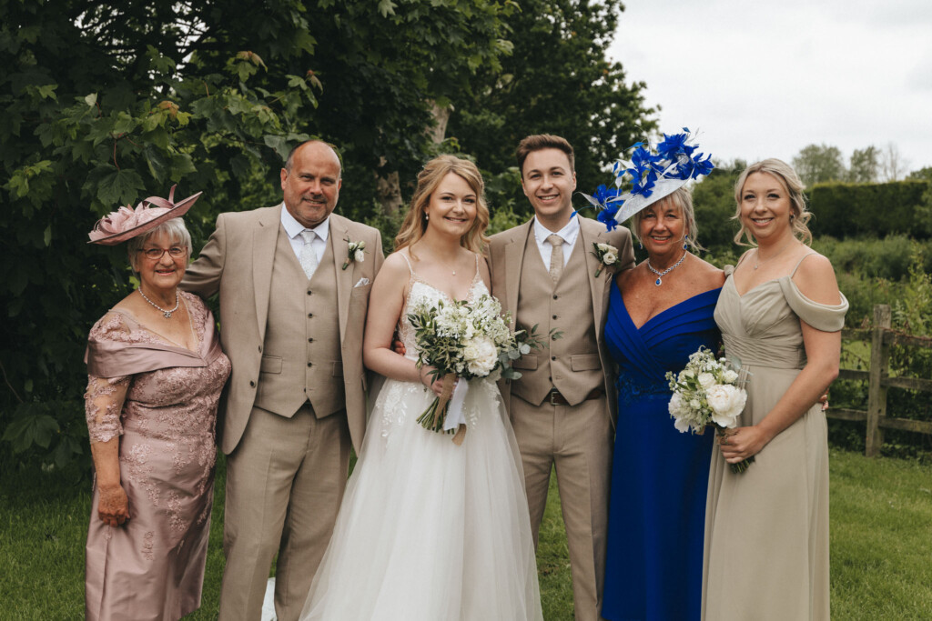 A wedding group photo featuring a bride in a white gown holding a bouquet, standing next to the groom in a beige suit. Flanking them are three women in elegant dresses, one wearing a blue fascinator, and a man in a beige suit. They are outdoors with greenery in the background. © Aimee Lince Photography