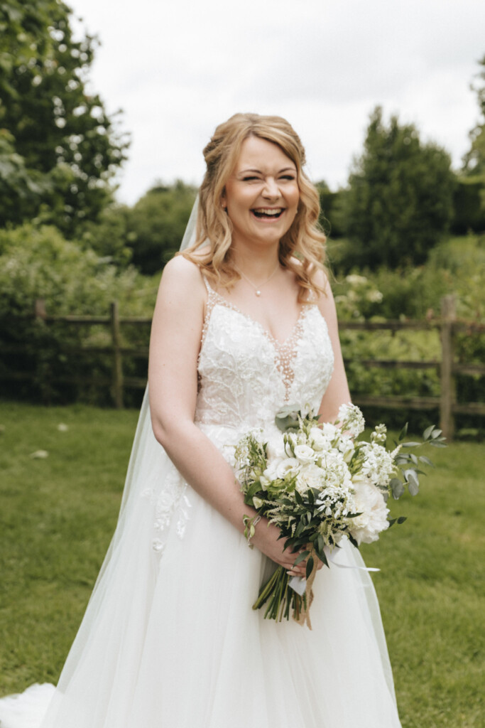 A smiling bride stands outdoors in front of greenery, wearing a white wedding dress with lace detailing. She holds a bouquet of white and green flowers. Her blonde hair is styled in loose curls. The background features a wooden fence and lush trees. © Aimee Lince Photography