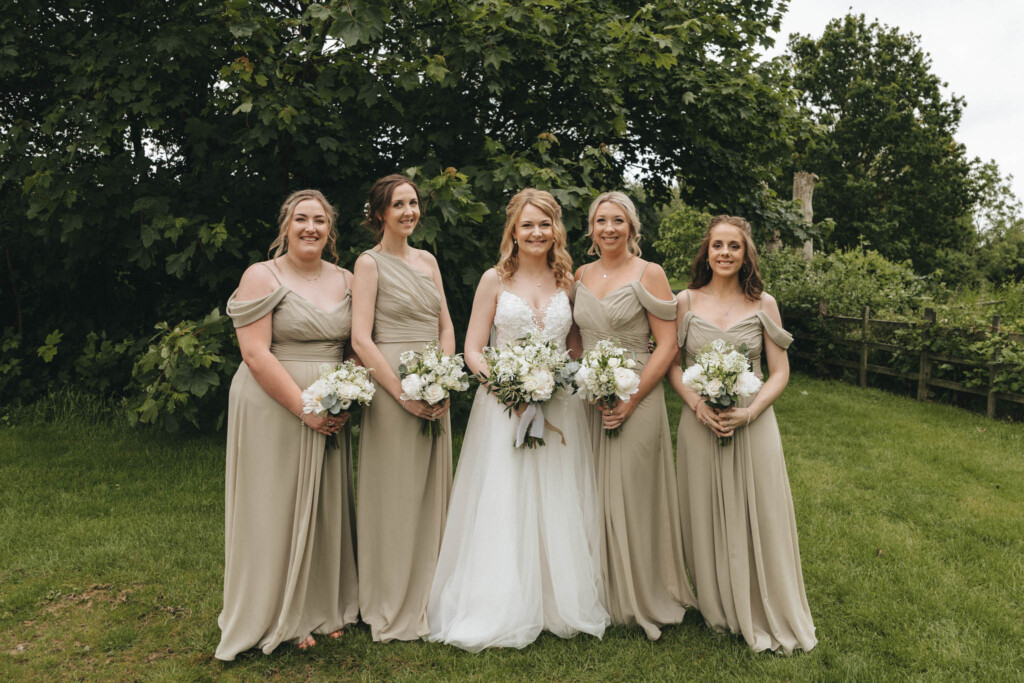 A bride in a white gown stands outside with four bridesmaids in matching light olive green dresses. They all hold bouquets of white flowers. The setting is a grassy area with lush green trees in the background, suggesting a serene, outdoor wedding scene. © Aimee Lince Photography