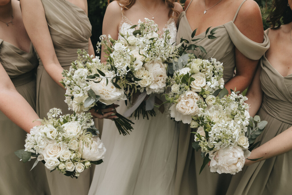 A group of women in sage green dresses hold large bouquets of white flowers with greenery. The bouquets feature roses, peonies, and other blossoms. The dresses have various styles, including off-the-shoulder and spaghetti straps. The women are standing closely together. © Aimee Lince Photography