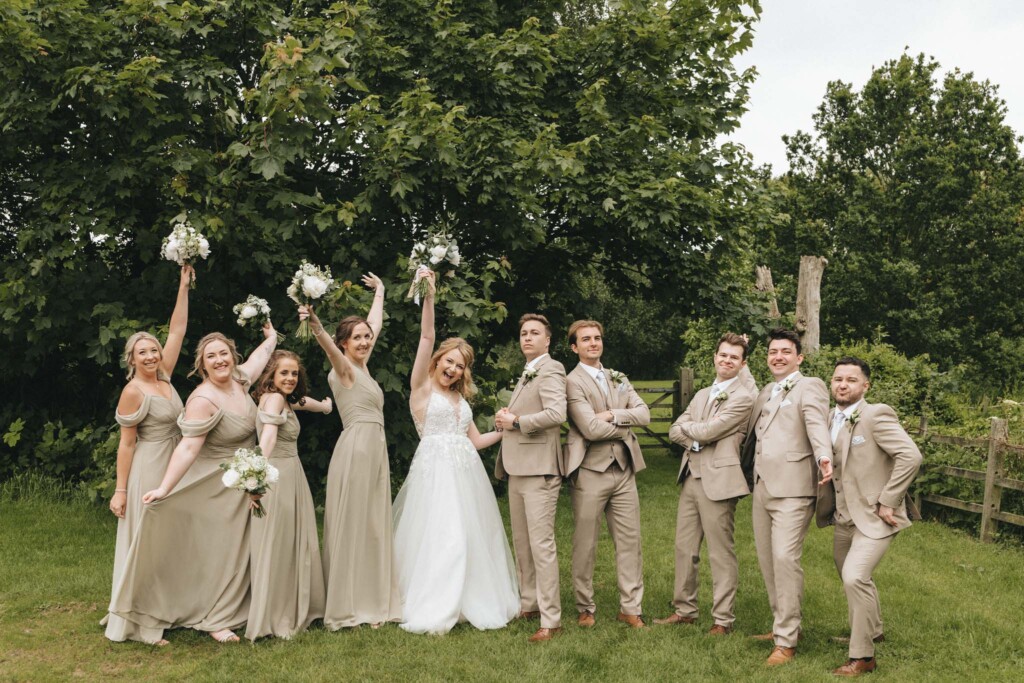 A wedding party poses outdoors in a garden. The bride, in a white gown, stands joyfully with her bridesmaids, who wear matching sage green dresses. The groom and groomsmen, in beige suits, stand confidently beside them. Lush trees and greenery serve as the backdrop. © Aimee Lince Photography