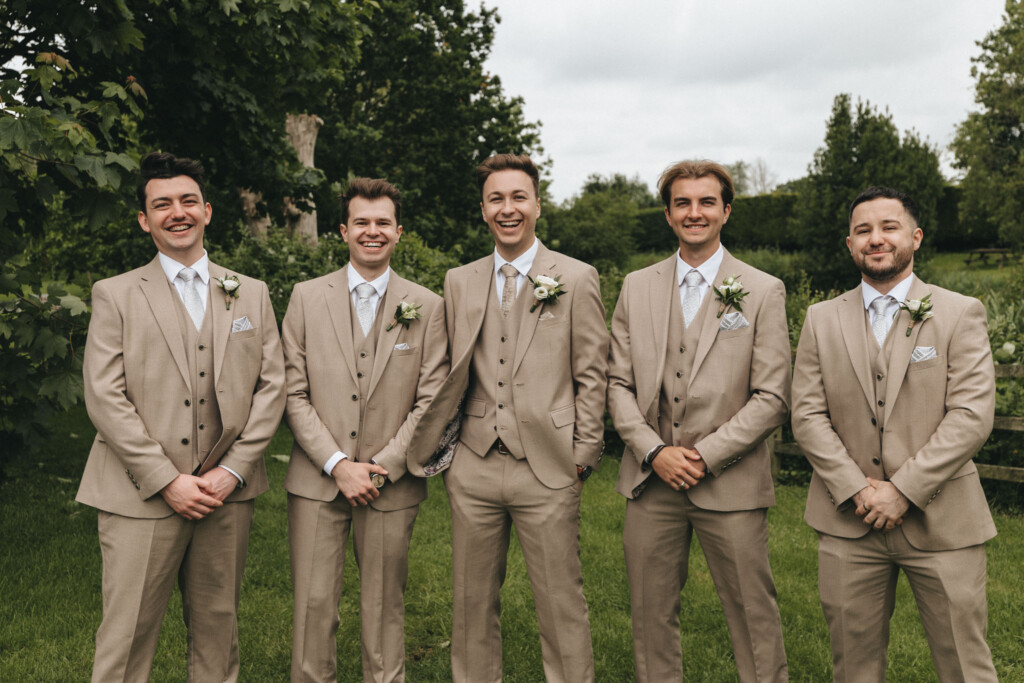 Five men in matching beige suits and vests stand in a row, smiling and posing with hands clasped or in pockets. Each has a boutonniere on their left lapel. They are outdoors on lush green grass with trees in the background under an overcast sky. © Aimee Lince Photography