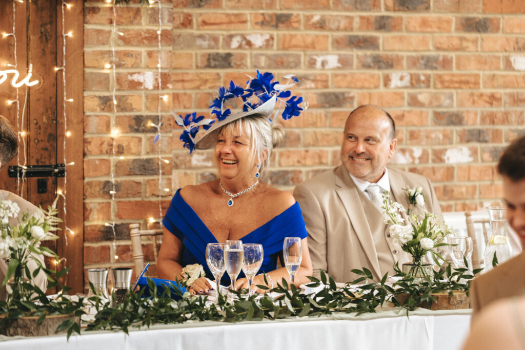 Hall Farm Hotel Wedding Photographer - Courtney & Tom © Aimee Lince Photography (496) An older couple sits at a wedding reception table. The woman wears a vivid blue dress and a matching hat adorned with blue flowers. The man wears a beige suit and tie. Both are smiling. The table is decorated with white flowers and greenery, and there is a brick wall in the background. © Aimee Lince Photography