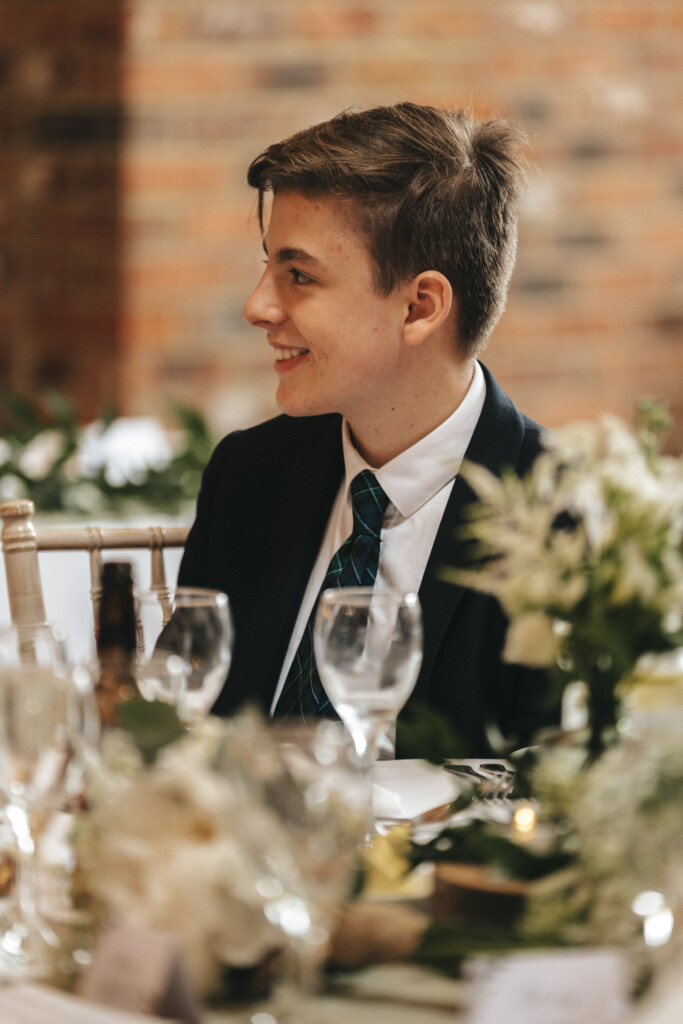 A young person in a suit and tie is seated at a dining table, smiling and looking to the side. The table is elegantly set with flowers, wine glasses, and a bottle, all against a background of a brick wall. © Aimee Lince Photography