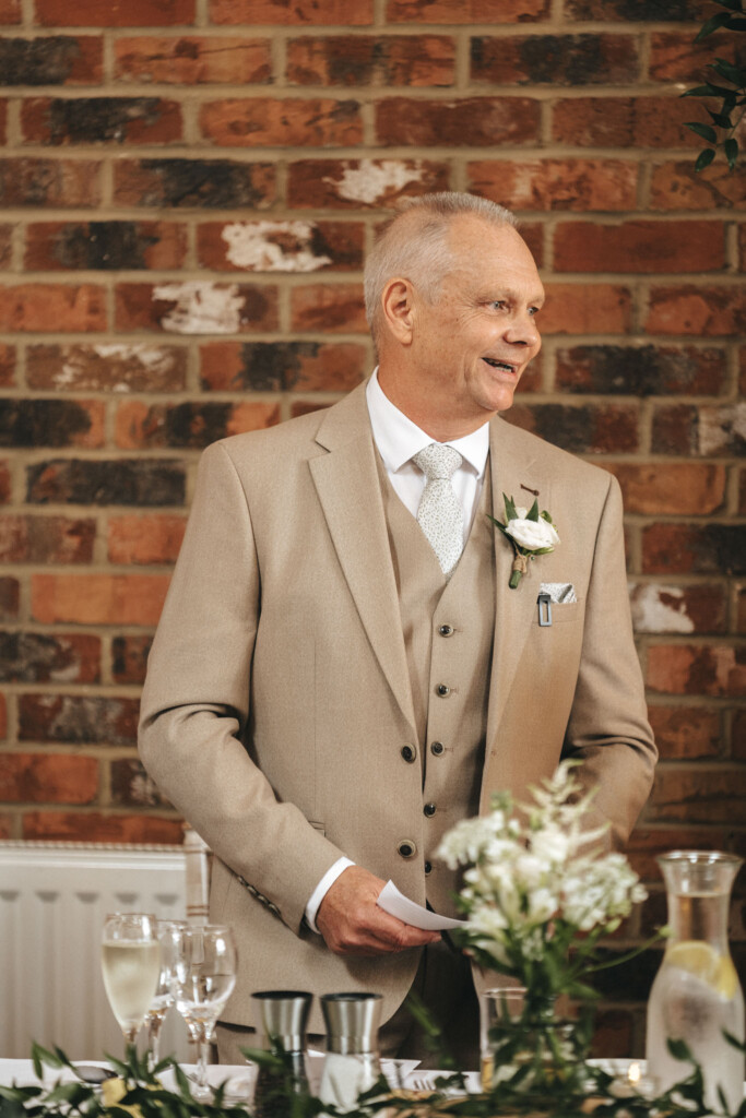 An older man in a beige suit and tie stands smiling in front of a brick wall. He holds a piece of paper and has a white flower boutonniere on his lapel. A table with flowers, glasses, and candles is in front of him. Greenery decorates the top right corner. © Aimee Lince Photography
