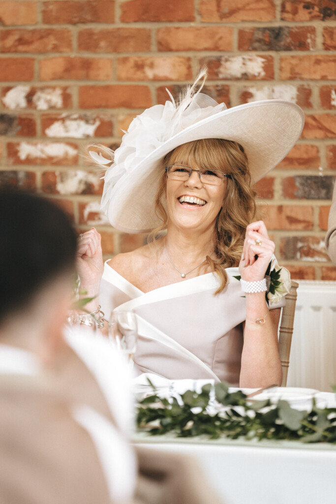 A woman wearing an elegant, off-the-shoulder cream dress and a large matching hat with feathers smiles joyfully while sitting at a table. She has glasses and wavy hair. A brick wall is in the background, and greenery decorates the table in front of her. © Aimee Lince Photography