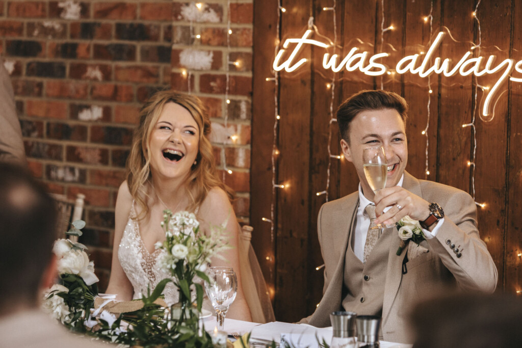 Hall Farm Hotel Wedding Photographer - Courtney & Tom © Aimee Lince Photography (514) A joyful couple sits at a wedding reception table. The woman, in a white dress, laughs openly, while the man, in a beige suit, raises a champagne flute, smiling. Behind them, a neon sign on a rustic brick wall partially reads "it was always." Greenery decorates the table. © Aimee Lince Photography