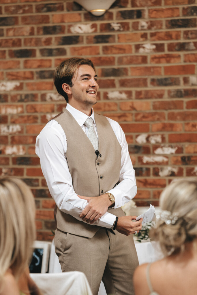 A man in a beige vest and white shirt smiles while standing in front of a brick wall. He is holding papers and appears to be speaking. Seated guests with blonde hair are in the foreground. A small light and part of a table with decorations are visible in the background. © Aimee Lince Photography