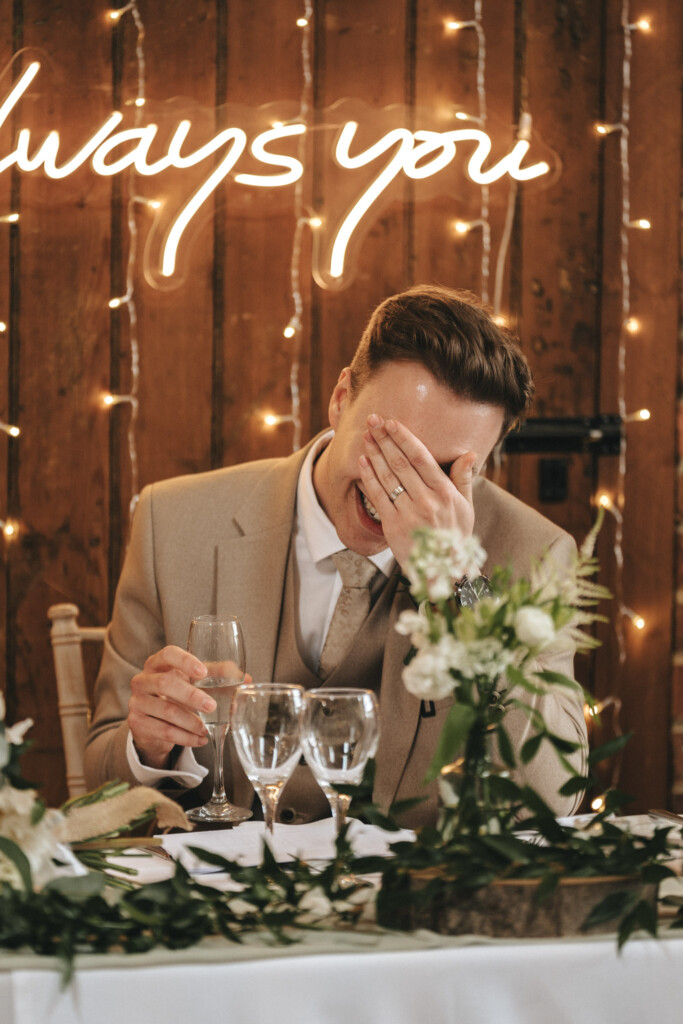 A man in a beige suit, seated at a decorated table with wine glasses and white flowers, laughs with his hand covering his face. Behind him, warm fairy lights and a glowing sign partially reading "always you" are visible against a wooden backdrop. © Aimee Lince Photography