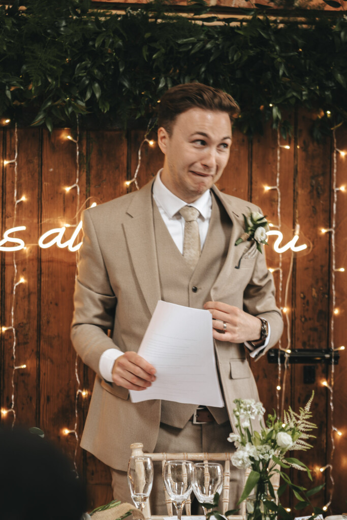 A man in a beige suit and tie stands smiling, holding papers, likely delivering a speech. Behind him is a wooden wall adorned with greenery and string lights. In front, a table holds champagne glasses and a floral arrangement. The atmosphere suggests a wedding or formal event. © Aimee Lince Photography
