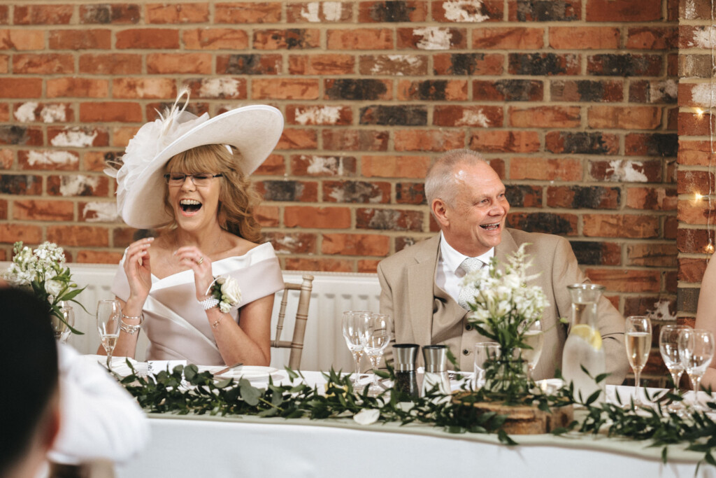 A smiling couple sits at a wedding reception table adorned with white flowers and greenery. The woman wears a large white hat and a white dress, while the man is dressed in a light beige suit. They are seated against a backdrop of rustic brick, enjoying the event. © Aimee Lince Photography