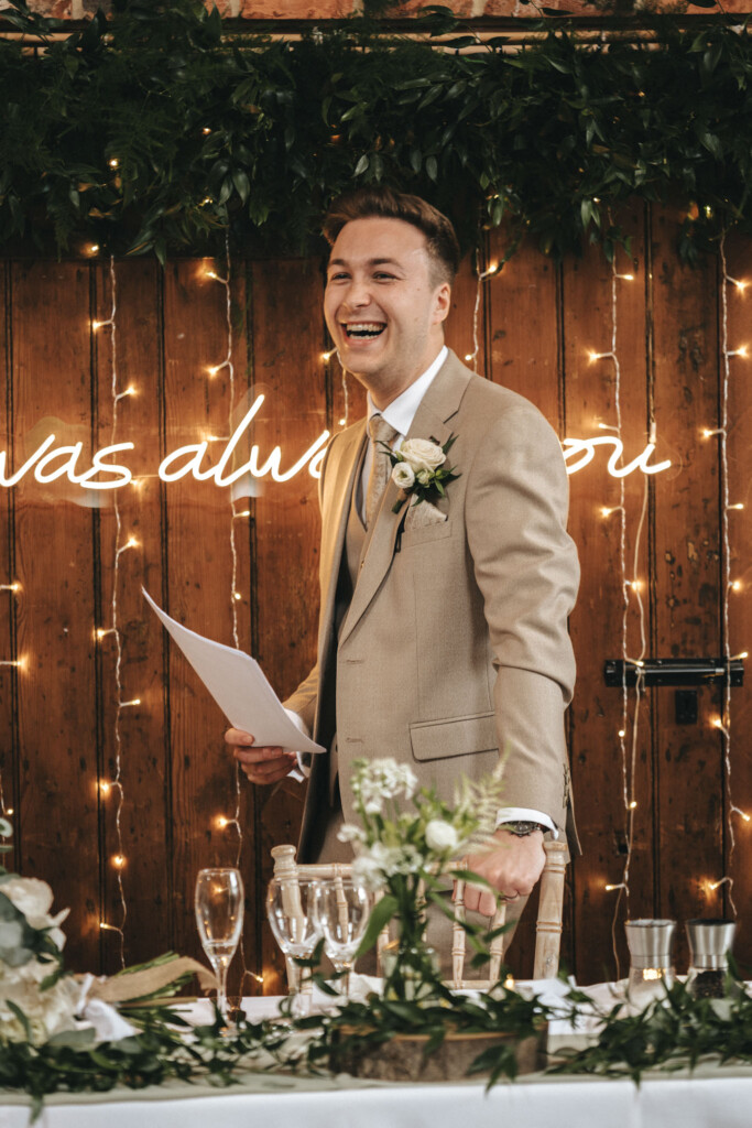 A man in a beige suit and tie, holding a piece of paper, stands and smiles. Behind him, a wooden wall is adorned with greenery and string lights. A partial neon sign glows softly. In the foreground, a table is set with floral arrangements and glassware. © Aimee Lince Photography