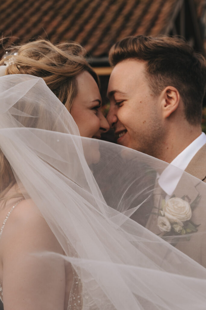A bride and groom stand close, smiling as their noses touch. The bride's veil billows around them, partially obscuring their faces. The groom wears a beige suit with a white rose boutonniere. They are outside, with a tiled roof visible in the background. © Aimee Lince Photography