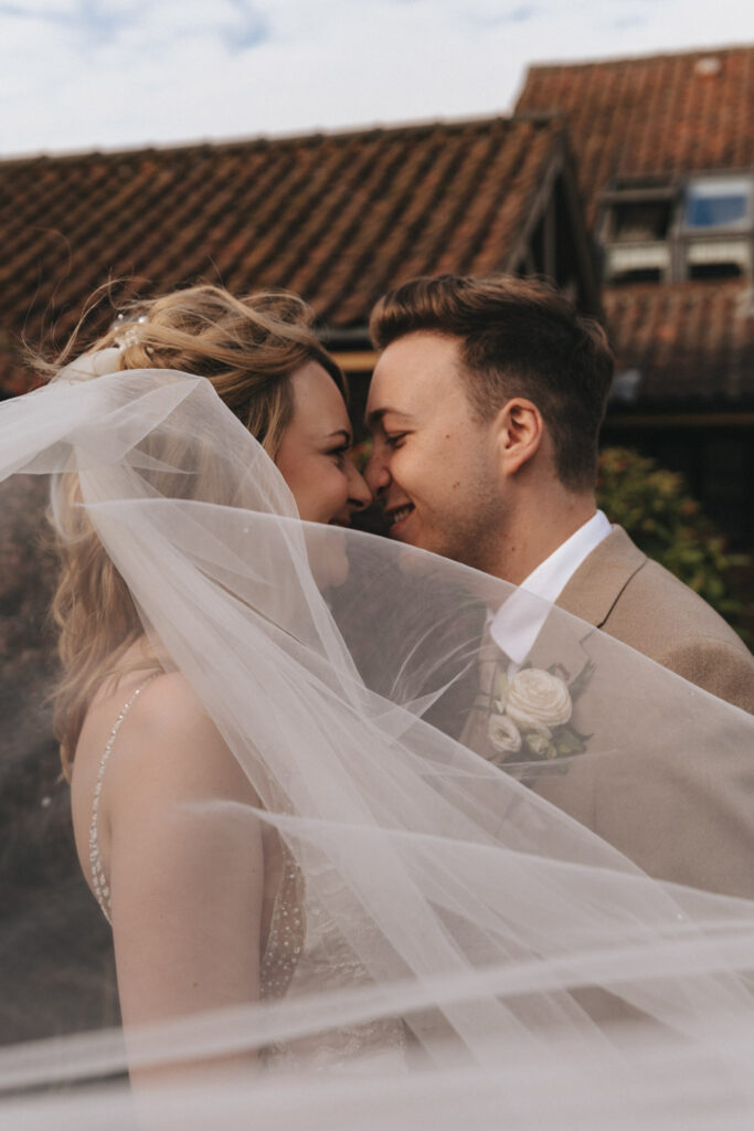 A bride and groom share an intimate moment, their foreheads touching and smiling warmly at each other. The bride's veil drapes in front of them, adding a soft focus. The groom wears a beige suit with a rose boutonniere. They stand against a building with a tiled roof in the background. © Aimee Lince Photography