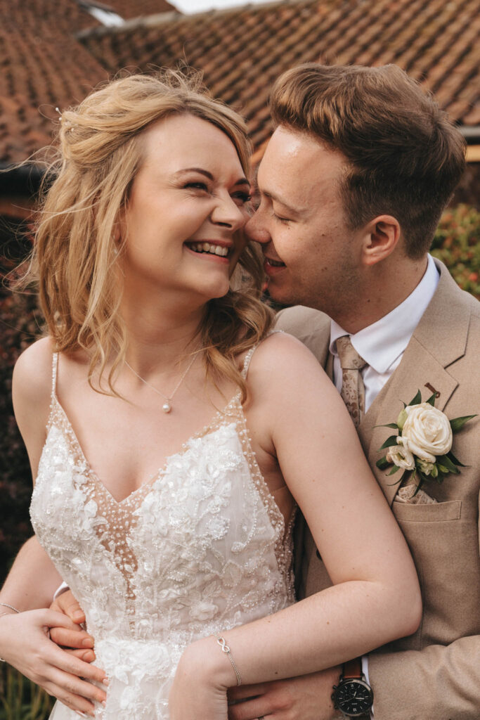 A smiling couple in wedding attire, with the bride in a lace dress and the groom in a beige suit with a white rose boutonniere. They are outdoors with a tiled roof and greenery in the background, embracing and looking into each other's eyes warmly. © Aimee Lince Photography