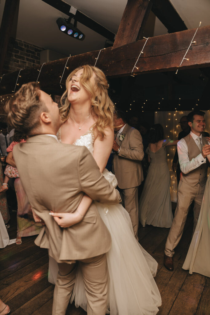 A couple dressed in tan formal wear dances joyfully on a wooden floor. The groom lifts the laughing bride, who wears a long white dress. They are surrounded by guests dressed in formal attire, with soft fairy lights and a rustic brick wall in the background, creating a warm, festive atmosphere. © Aimee Lince Photography
