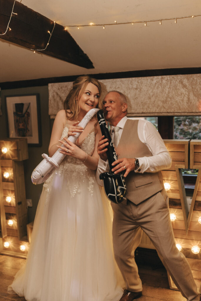 A bride in a white dress playfully holds an inflatable saxophone, smiling at a man in a tan vest and pants who pretends to play a black inflatable saxophone. They're standing in a warmly lit room with string lights and a large, lit letter display in the background. © Aimee Lince Photography - Wedding photographer in Lincolnshire, Yorkshire & Nottinghamshire