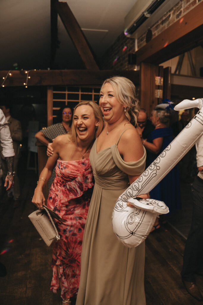 Two women are laughing and embracing at a party. One wears a strapless pink floral dress and holds a clutch. The other wears an off-shoulder beige gown and holds a white inflatable saxophone. The background features people, wooden beams, and string lights. © Aimee Lince Photography