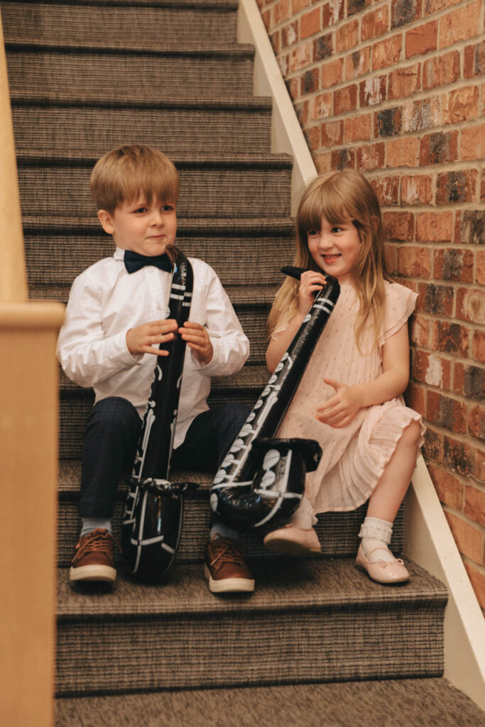 Two young children sitting on carpeted stairs, each holding an inflatable saxophone. The boy, wearing a white shirt and trousers, and the girl, in a light pink dress, are smiling. The wall beside them is red brick. They appear to be playful and happy. © Aimee Lince Photography